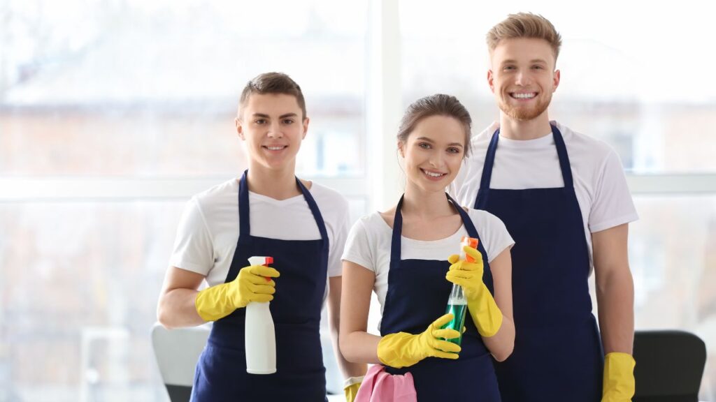 three people, one woman and two men, wearing cleaning gear and holding cleaning products while smiling
